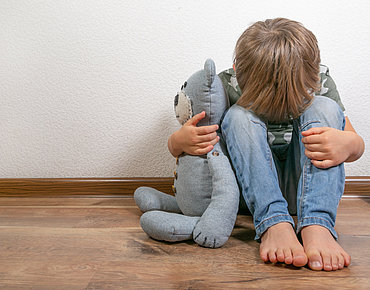 Kind sitzt verängstlich mit seinem Teddy an der Wand